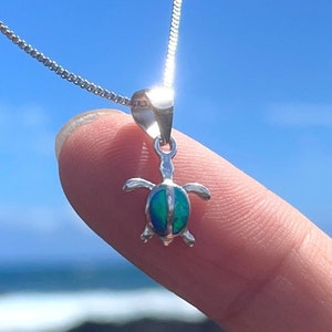 Op de afbeelding: Een zilveren ketting met een klein, groen opaal schildpad hanger. De hanger is op een vinger tegen een blauwe lucht en oceaan achtergrond.