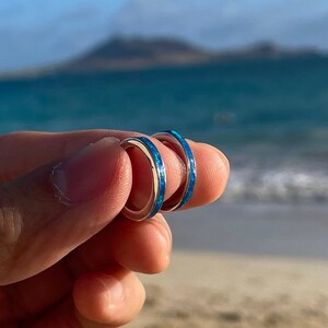 May include: Two thin silver rings with a blue inlay. The rings are held in a hand against a background of a beach and ocean.