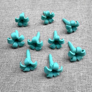 May include: Nine turquoise-colored, miniature succulent-shaped polymer clay charms are arranged on a gray textured surface. Each charm has a central point with curved petals, resembling a small flower or succulent plant. The charms are evenly spaced.