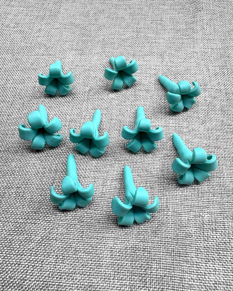 May include: Nine turquoise-colored, miniature succulent-shaped polymer clay charms are arranged on a gray textured surface. Each charm has a central point with curved petals, resembling a small flower or succulent plant. The charms are evenly spaced.
