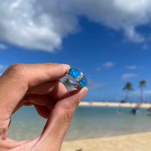 Puede incluir: Un anillo de plata con una piedra de &oacute;palo azul engastada en una banda ancha. El anillo se muestra sobre un fondo de playa y oc&eacute;ano.