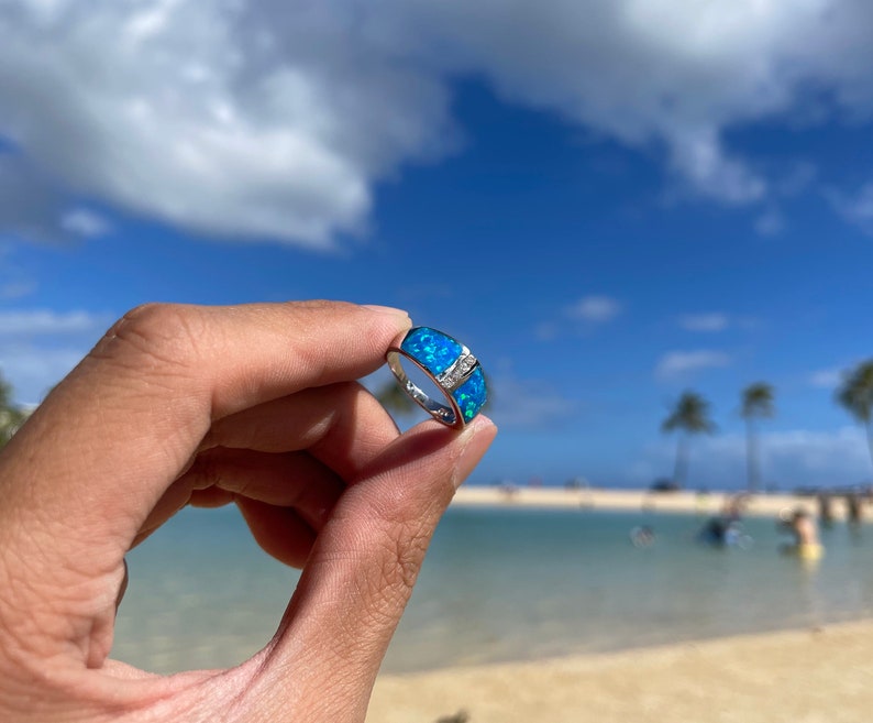 Puede incluir: Un anillo de plata con una piedra de &oacute;palo azul engastada en una banda ancha. El anillo se muestra sobre un fondo de playa y oc&eacute;ano.