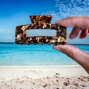 Puede incluir: Una pinza para el pelo de color marrón y dorado de caparazón de tortuga. La pinza tiene una forma rectangular con una superficie lisa y plana y una fila de dientes en la parte inferior.