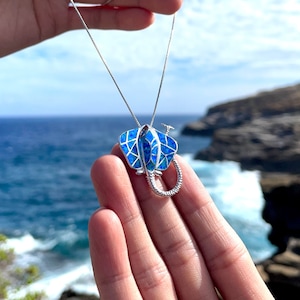 Op de afbeelding: Een zilveren ketting met een blauwe opaal-roggenhanger. De hanger is gedetailleerd met een textuur, geometrisch patroon. De ketting is gefotografeerd tegen een achtergrond van blauw water en een rotsachtige kust.