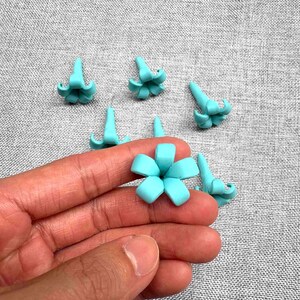 May include: A hand holding a light blue flower-shaped craft item, surrounded by several similar items. The items are small, with a detailed petal design. The background is a textured gray fabric.