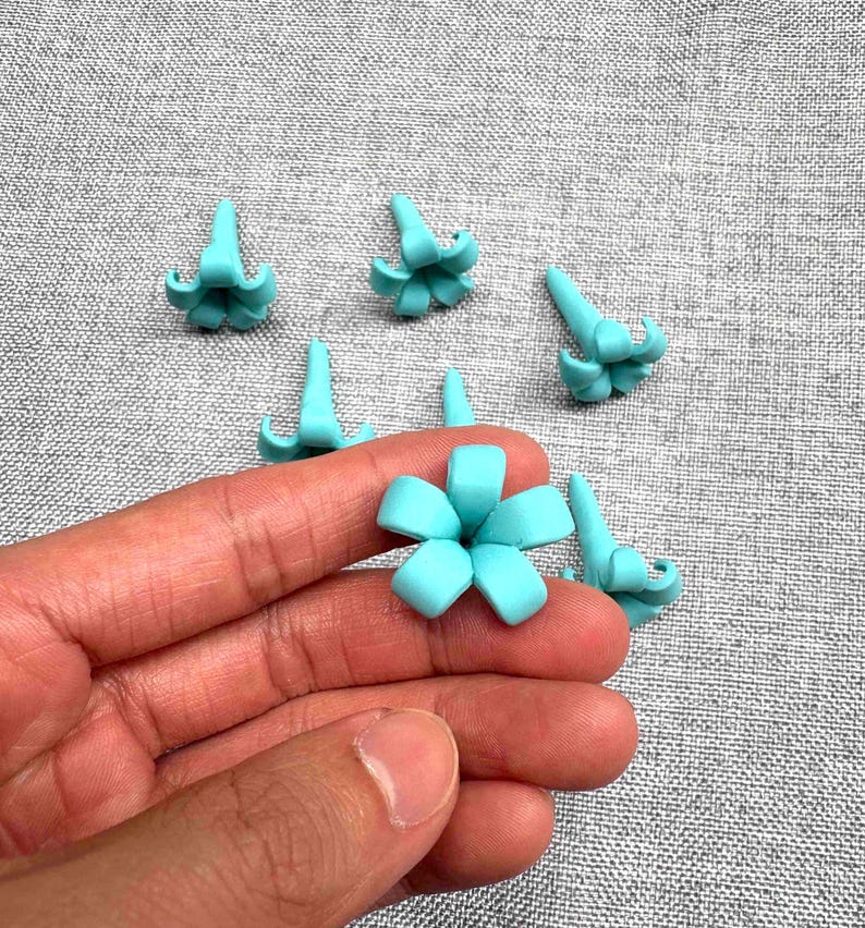 May include: A hand holding a light blue flower-shaped craft item, surrounded by several similar items. The items are small, with a detailed petal design. The background is a textured gray fabric.