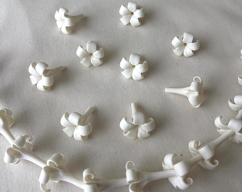 May include: A collection of small, white, flower-shaped beads and a floral vine. The beads are scattered across a light-colored surface. The flowers have a delicate, layered petal design. The vine is also white, with similar flower buds attached.