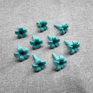 May include: A collection of nine small, turquoise-colored, flower-shaped embellishments. Each flower has a central cone shape with curved petals. The objects are arranged on a gray, textured surface.