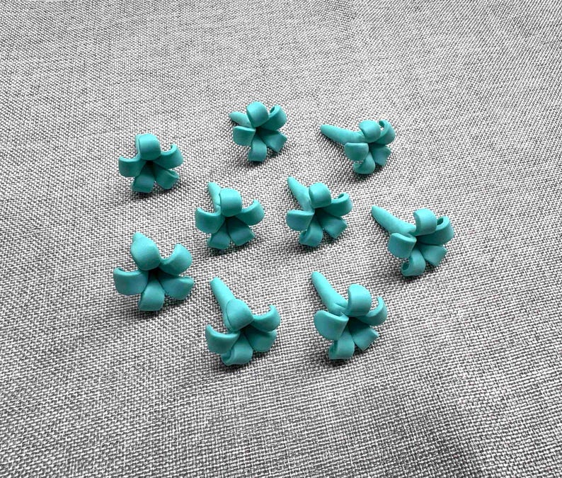 May include: A collection of nine small, turquoise-colored, flower-shaped embellishments. Each flower has a central cone shape with curved petals. The objects are arranged on a gray, textured surface.