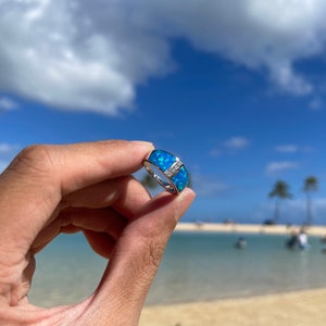 Puede incluir: Un anillo de plata con una piedra de &oacute;palo azul engastada en una banda. El anillo est&aacute; sostenido por una mano sobre un fondo de cielo azul y playa.