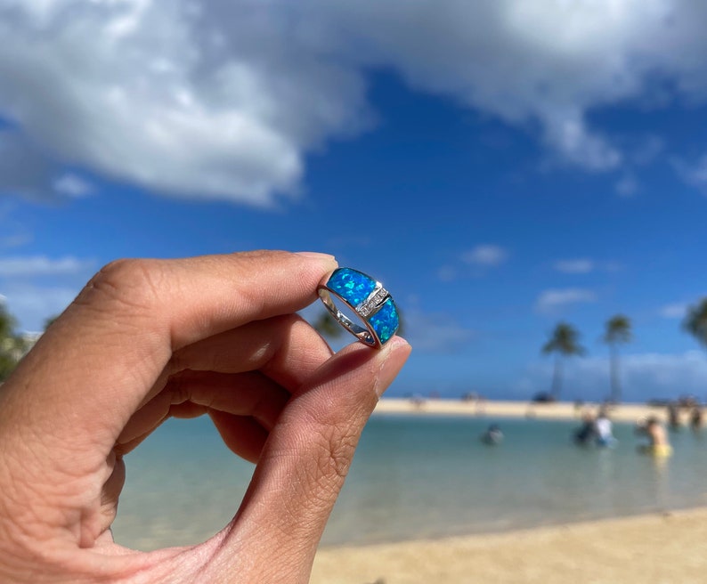 Puede incluir: Un anillo de plata con una piedra de &oacute;palo azul engastada en una banda. El anillo est&aacute; sostenido por una mano sobre un fondo de cielo azul y playa.