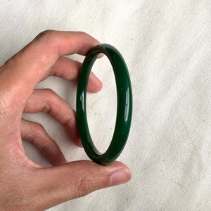 May include: A glossy, dark green bangle bracelet. The circular bracelet is held against a white fabric background. The bangle is a solid color and appears to be made of a smooth material.