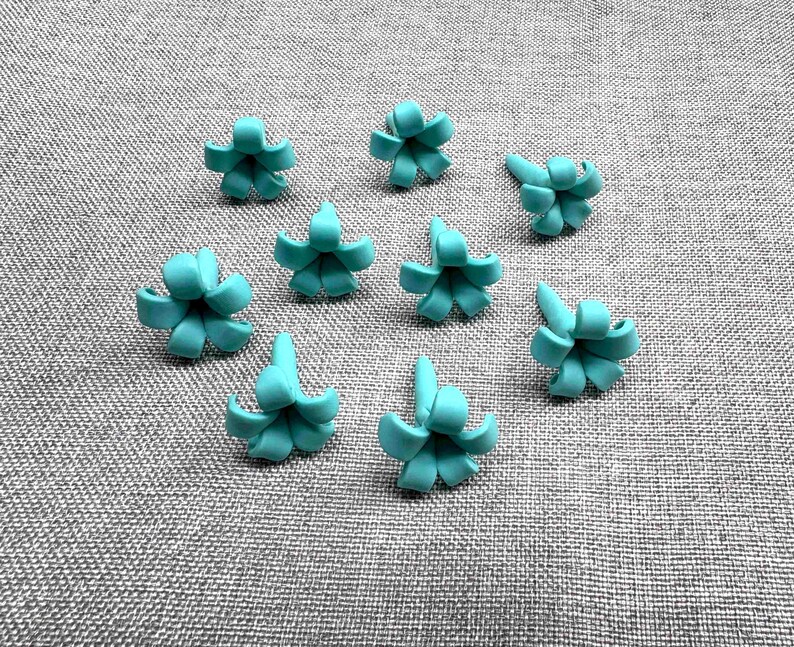 May include: Nine turquoise flower-shaped beads are arranged on a gray textured surface. Each bead has a central point with curved petals, resembling small blossoms. The beads are evenly spaced, creating a visually appealing pattern.