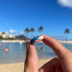 May include: A silver ring with a multi-colored opal inlay. The opal has shades of blue, pink, and white. The ring is set against a background of a beach and ocean.