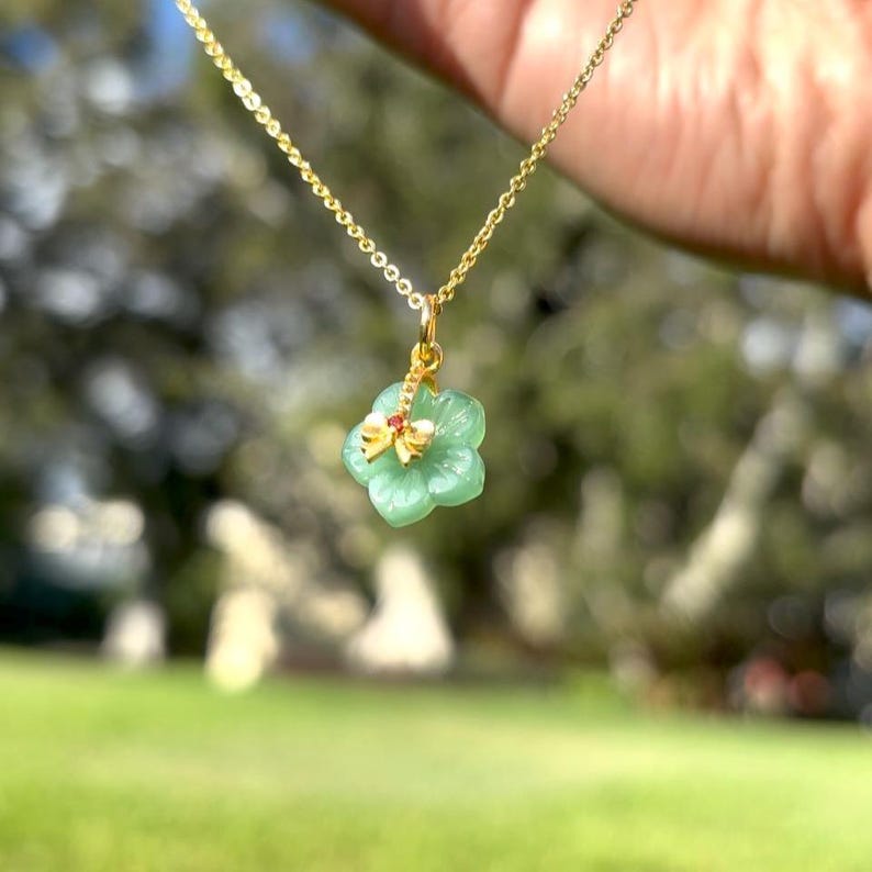 K&ouml;nnte beinhalten: Eine goldene Halskette mit einem gr&uuml;nen Blumenanh&auml;nger. Der Blumenanh&auml;nger hat einen goldenen Libellen-Charm mit roten Akzenten. Der Hintergrund ist eine verschwommene Ansicht von gr&uuml;nem Gras und B&auml;umen.