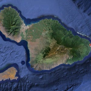May include: Satellite image of a lush green island with mountainous terrain, surrounded by deep blue ocean. The island has a rugged coastline and a small, adjacent island. The word "Hana" is labeled with a red marker on the eastern side of the island.