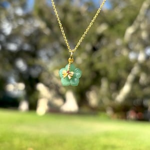K&ouml;nnte beinhalten: Eine goldene Halskette mit einem Anh&auml;nger in Form einer gr&uuml;nen Blume, verziert mit einer goldenen Schleife und einem kleinen roten Edelstein. Die Halskette vor einem verschwommenen gr&uuml;nen Hintergrund.