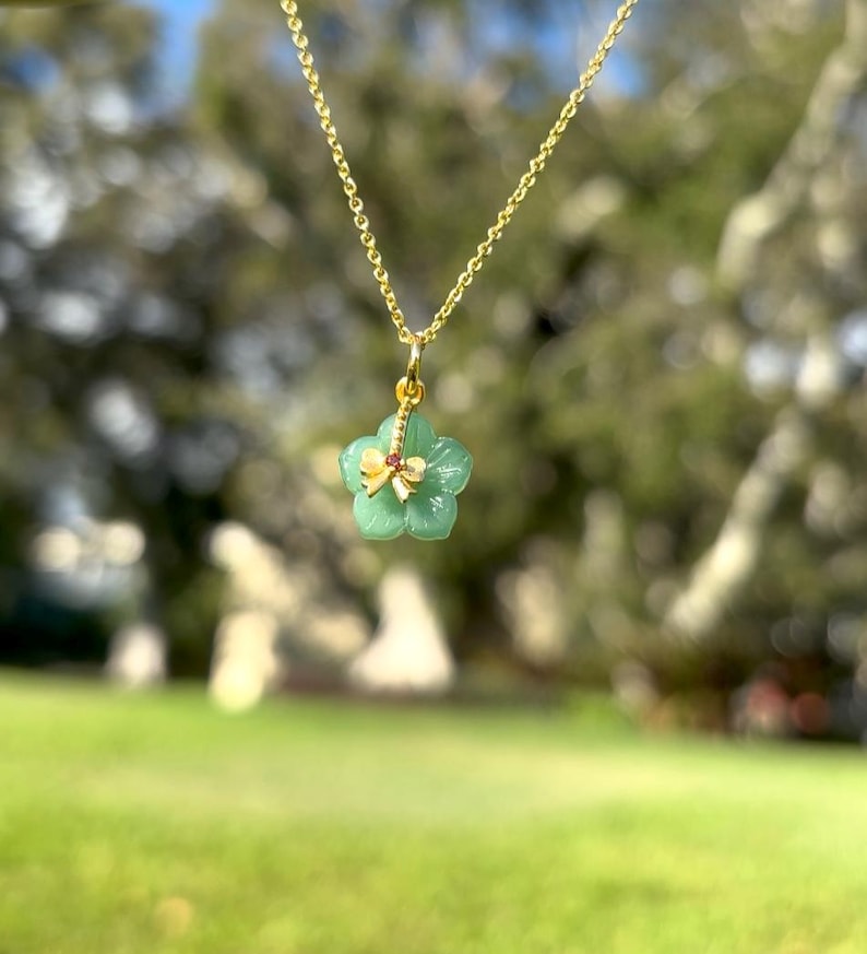 K&ouml;nnte beinhalten: Eine goldene Halskette mit einem Anh&auml;nger in Form einer gr&uuml;nen Blume, verziert mit einer goldenen Schleife und einem kleinen roten Edelstein. Die Halskette vor einem verschwommenen gr&uuml;nen Hintergrund.