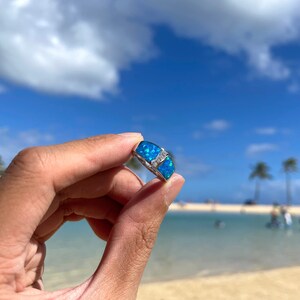 Puede incluir: Un anillo de plata con una piedra de &oacute;palo azul engastada en una banda de plata. El anillo se sostiene en una mano contra un fondo de playa y oc&eacute;ano.