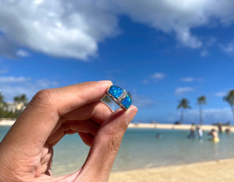 Puede incluir: Un anillo de plata con una piedra de &oacute;palo azul engastada en una banda de plata. El anillo se sostiene en una mano contra un fondo de playa y oc&eacute;ano.