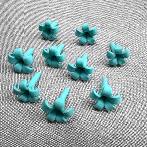 May include: Nine turquoise flower-shaped craft embellishments are scattered on a gray textured surface. Each flower has a central point with curved petals. The embellishments are evenly spaced and appear to be made of a smooth material.