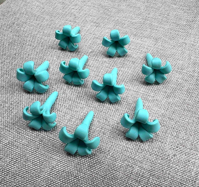 May include: Nine turquoise flower-shaped craft embellishments are scattered on a gray textured surface. Each flower has a central point with curved petals. The embellishments are evenly spaced and appear to be made of a smooth material.