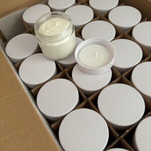 Pode incluir: Uma caixa cheia de recipientes redondos brancos. Um recipiente está aberto e mostra um creme branco dentro.