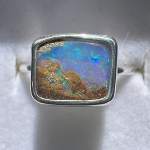 May include: A silver ring featuring a square-shaped opal gemstone set in a bezel setting. The opal displays blue, green, and brown hues.