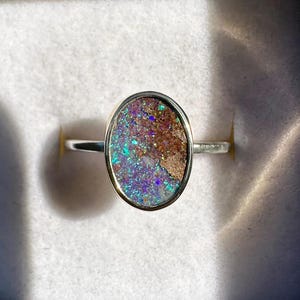 May include: Oval opal ring with a silver band, presented in a black box with a white interior. The opal displays a mix of brown, blue, and purple hues. The ring is shown in natural light.