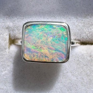 May include: A silver ring featuring a square-shaped opal gemstone. The opal has a white base with green, blue, and orange iridescent flashes.