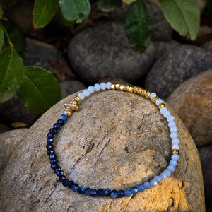 May include: A blue and white ombre beaded bracelet with a gold star charm. The bracelet is made of small, round beads that are graduated in size from small to large. The beads are strung on a clear elastic cord.