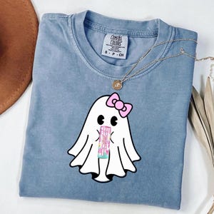 Op de afbeelding: Een lichtblauw Comfort Colors T-shirt met een cartoon spookontwerp. Het spook heeft een roze strik en houdt een roze en wit object vast. Een gouden ketting met een hanger ligt op het T-shirt. Het T-shirt heeft een "Comfort Colors" label.