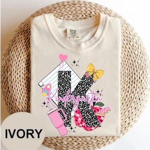 May include: Ivory-colored t-shirt with a colorful graphic design for kindergarten. The design includes a large letter "K" with a black and white pattern, a pink tumbler, a yellow bow, and the word "Kindergarten".