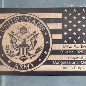 May include: A wooden plaque with a black and white American flag design. The plaque features the United States Army seal and the text "UNITED STATES ARMY". The plaque also includes the text "MAJ Audie Murphy, 20 June 1925 - 28 May 1971, recipient of the Congressional Medal of Honor and 3 Purple Hearts."
