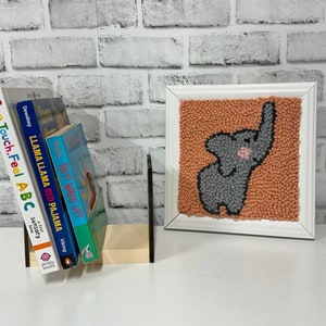 Peut inclure: Une œuvre d'art en pointillé encadré représentant un éléphant gris avec un nez rose sur un fond pêche. L'œuvre d'art est sur une étagère blanche avec une pile de livres pour enfants.
