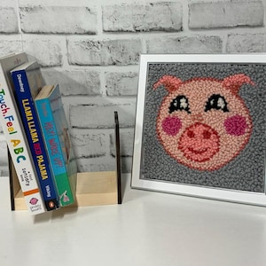 Peut inclure: Une œuvre d'art en point d'aiguille encadrée représentant un cochon rose avec un visage souriant. Le cochon a des yeux noirs et des joues roses. L'arrière-plan est en feutre gris.