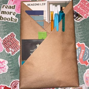Puede incluir: Un organizador de bolsillo de papel marrón con una lista de lectura, bolígrafos y marcadores. El organizador está rodeado de pegatinas con frases relacionadas con los libros como "Read More Books" y "Hello, I'm addicted to buying books."