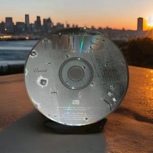 Puede incluir: Un CD plateado con el texto "MMS MUSIC IS MY SAVIOR" y el logotipo de Capitol Records. El disco tiene una etiqueta de advertencia para padres. El fondo muestra una silueta de paisaje urbano contra un atardecer.