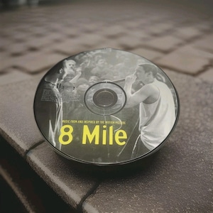 Puede incluir: Un disco compacto de la película "8 Mile" con una imagen en blanco y negro de un hombre cantando. El disco tiene texto amarillo que dice "8 Mile" y el texto "MUSIC FROM AND INSPIRED BY THE MOTION PICTURE".