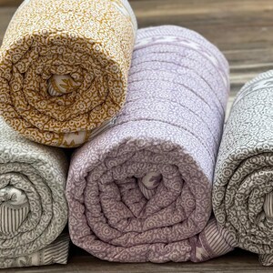 May include: Four rolled-up quilts in gold, lavender, and grey. Each quilt has a repeating floral pattern. The quilts are tightly rolled, showing the patterned fabric and the contrasting striped edges. The quilts are likely for bedding or home decor.