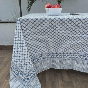 May include: A white tablecloth with a blue and gray geometric pattern and floral border. A white bowl filled with red apples sits on the table. The tablecloth is draped over a wooden table.