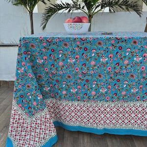 May include: A turquoise tablecloth featuring a floral design in pink and maroon. The tablecloth has a white and red geometric border and a solid turquoise edge. A white bowl of red apples sits on the table.
