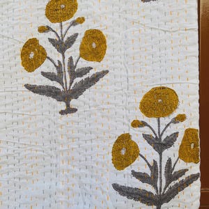 Puede incluir: Textil blanco con un estampado floral repetido. Las flores son amarillas con tallos y hojas grises. La tela tiene una textura acolchada con costuras amarillas y grises.