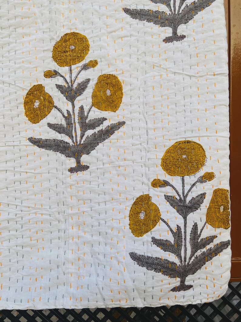 Puede incluir: Textil blanco con un estampado floral repetido. Las flores son amarillas con tallos y hojas grises. La tela tiene una textura acolchada con costuras amarillas y grises.