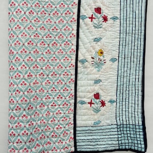 Könnte beinhalten: Eine Steppdecke mit floralem Muster in Rot und Türkis auf weißem Grund. Das Design umfasst ein zentrales Feld mit floralen Motiven und einen gestreiften Rand. Die Kanten sind mit einer dunkelblauen Paspel versehen.