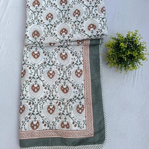May include: A folded quilt featuring a white background with a repeating pattern of brown and dark green floral motifs. The quilt is bordered by a thin brown line and a wider dark green edge, finished with a small striped trim along the outer border.