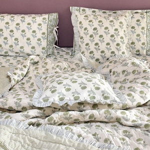 May include: A bed set with a white duvet cover, pillows, and a striped blanket. The bedding has a repeating pattern of green floral designs. The pillows and duvet cover have ruffled edges. The background is a solid purple.