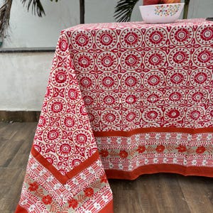 Può includere: Una tovaglia rettangolare caratterizzata da un motivo geometrico circolare ripetuto in rosso e bianco sulla superficie superiore, con un ampio bordo arancione e una fascia decorativa di motivi floreali arancioni e strisce lineari.