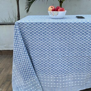 Puede incluir: Un mantel de mesa con estampado azul y blanco con un borde blanco. El mantel está colocado sobre un suelo de madera con un cuenco de manzanas rojas encima.