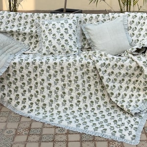May include: A white floral patterned quilt and matching pillows with ruffled edges are displayed on a sofa. The quilt and pillows feature a repeating pattern of green flowers. A striped pillow is also included.